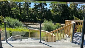 Eine Holzterrasse mit Glasumrandung, die zu einer Holztreppe führt und einen Blick auf eine natürliche Landschaft mit Bäumen und einem Teich bietet.