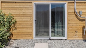 Fachada de madera con puerta corredera de vidrio, un pequeño espacio exterior con grava y una manguera a la derecha.