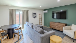 Una sala de estar moderna con un sofá seccional gris, una mesa de comedor de madera con sillas, una mesita redonda pequeña, un televisor montado en la pared y un letrero que dice "SALT LAKE CITY".