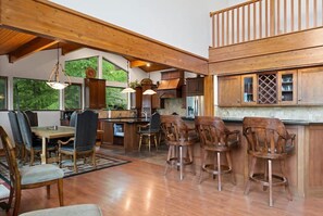 A spacious kitchen with wooden cabinets and a large island, a dining area with a table and chairs, and a view of greenery through large windows.
