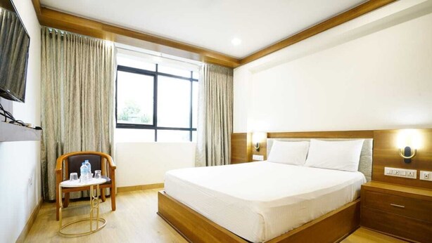 A hotel room with a large bed, wooden headboard, a bedside table, a chair, a small table with glasses and a bottle, and a window with curtains. - Hotel Astro Marine (Kanayannur)