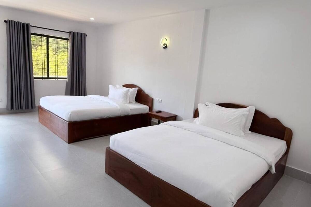 Una camera d'albergo con due letti, un'ampia finestra con tende, un comodino con una lampada e una luce a parete.