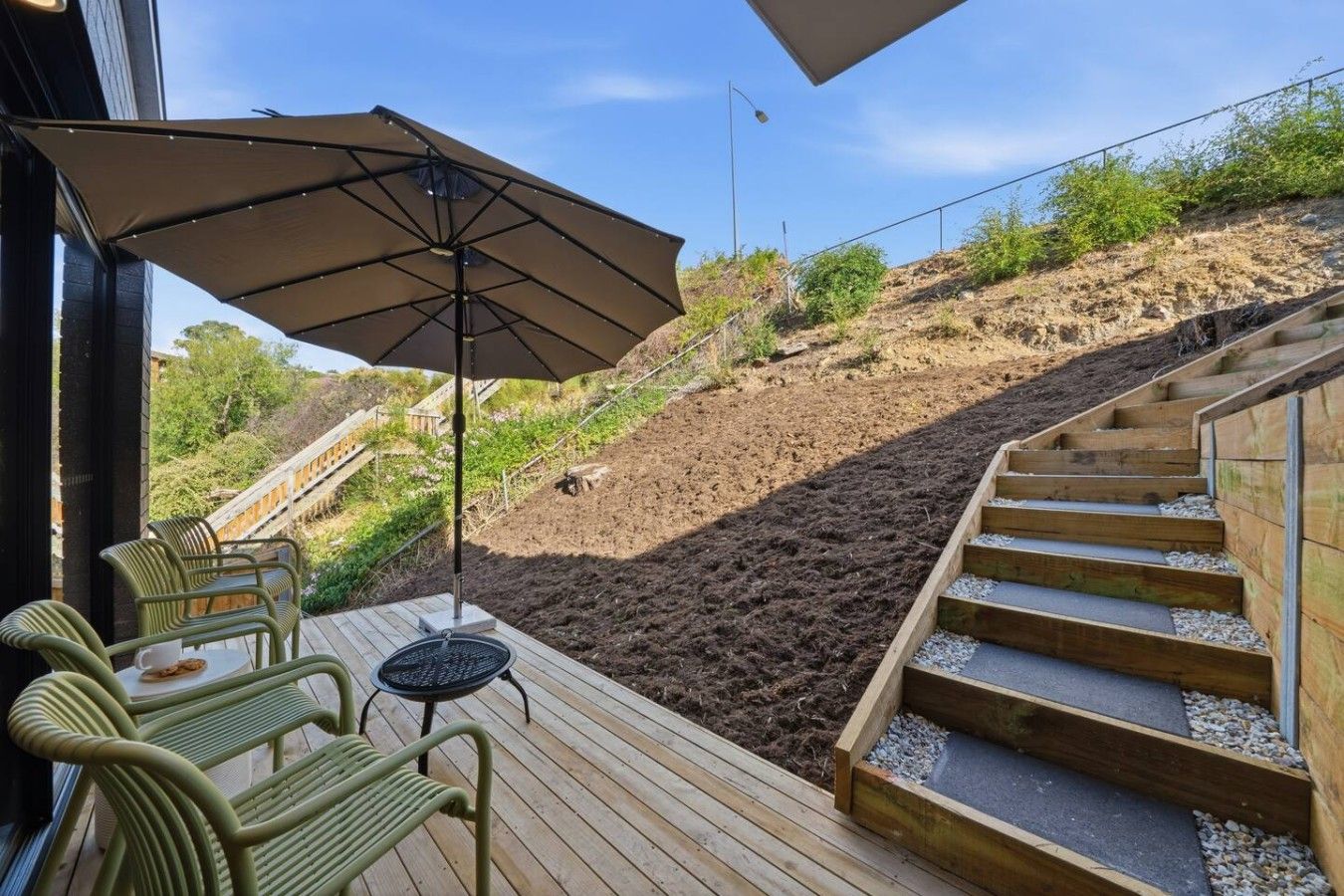 A deck with outdoor furniture, a large umbrella, and a view of a landscaped hillside.