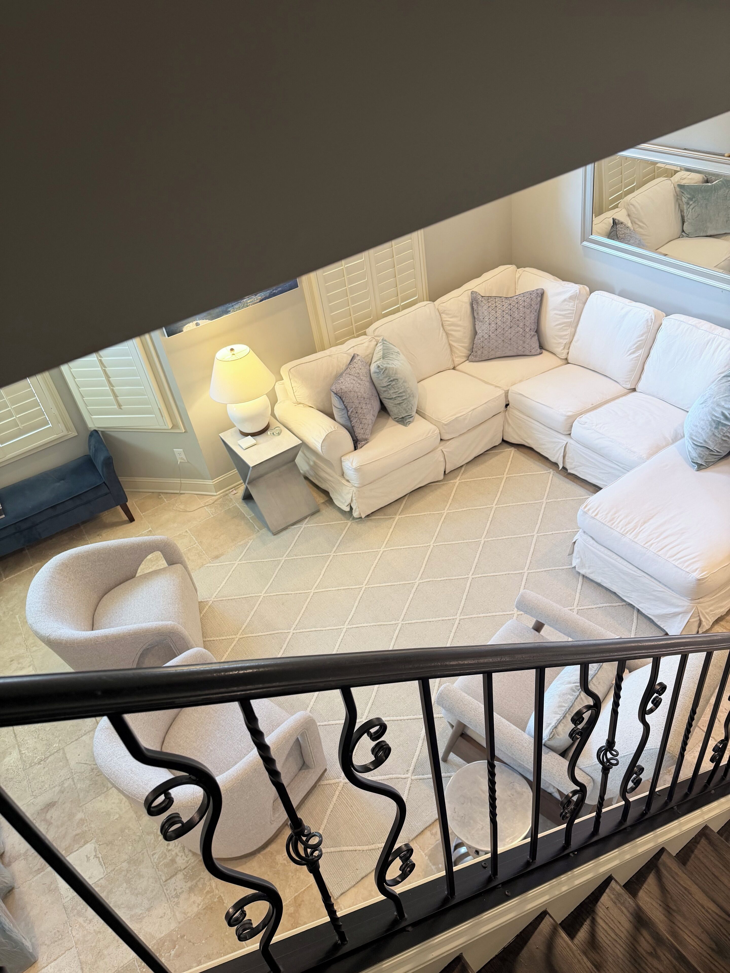 A modern living room with a white sectional sofa, a small round table with a lamp, and a grey armchair. A staircase with a black railing is visible in the foreground.