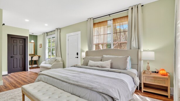 A bedroom with a large bed, a bedside table with a lamp, a bench, and a window with curtains. - Magnolia Soul- Modern Southern Retreat (Macon)