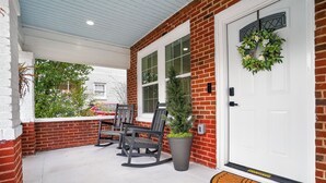 A covered porch with a brick wall, a white door with a wreath, and a potted plant. - Magnolia Soul- Modern Southern Retreat (Macon)