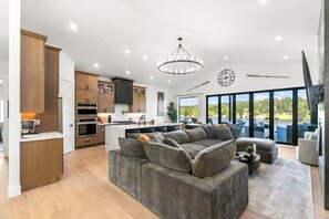 A spacious living area with a large gray sectional sofa, a kitchen with wooden cabinets, and a view of the outdoors through large windows. - Luxe Port Lookout with waterfront with dock, theater, hot tub, and huge deck (Kitsap County)