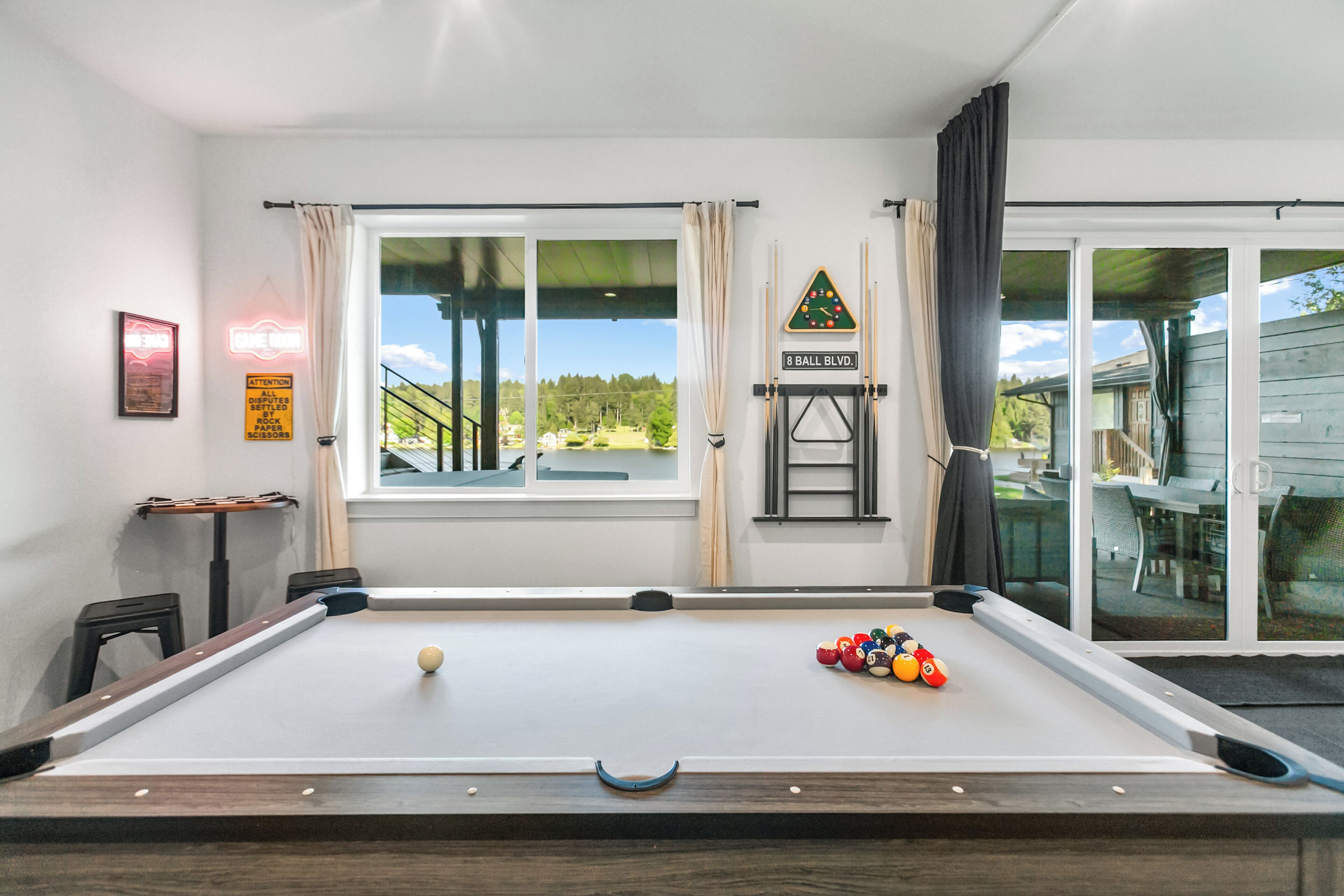 Een pooltafel met ballen, een raam met gordijnen, een neonbord, en uitzicht op een huis en bomen.