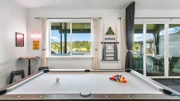 Een pooltafel met ballen, een raam met gordijnen, een neonbord, en uitzicht op een huis en bomen.