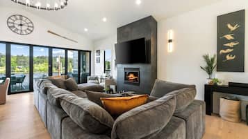 Ein modernes Wohnzimmer mit einem großen, grauen Ecksofa, einem Kamin und einem großen Fenster mit Blick auf Grünflächen.