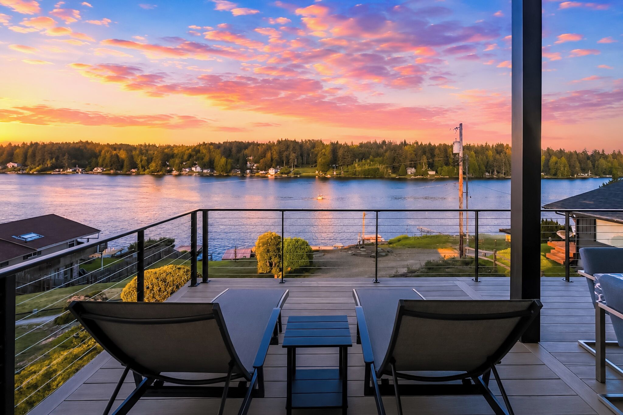 Un balcón con dos sillas y una mesa, con vista a un lago bordeado por un bosque y un cielo al atardecer.