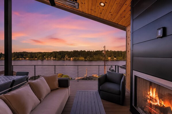 En træterrasse med sofa og stole, en pejs og udsigt over en sø i solnedgangen.