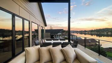 A modern outdoor seating area with a sofa and chairs, large windows, and a view of a lake and hills.