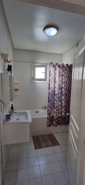 A bathroom with a white sink, a bathtub, a shower curtain, a window, and a ceiling light. - Spacious apartment, perfect for families and guests (Aouina)