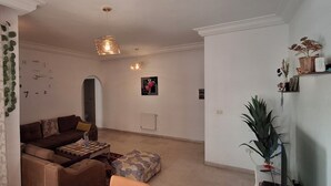 Ein Wohnzimmer mit einer Couch, einem Couchtisch und einem gemusterten Teppich. Rechts steht eine Pflanze und an der Wand hängt eine Uhr.