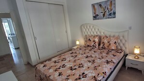 Ein Schlafzimmer mit einer blumigen Tagesdecke, einem gepolsterten Kopfteil, zwei Nachttischlampen und einem an der Wand hängenden Kunstwerk.