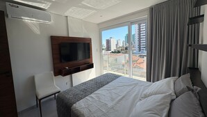 Habitación de hotel con cama, televisor montado en la pared, una silla y balcón con vista a la ciudad.