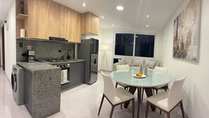 Una cocina moderna con encimeras de granito, horno empotrado y lavavajillas. Cuenta con un comedor con una mesa redonda de vidrio y sillas blancas.