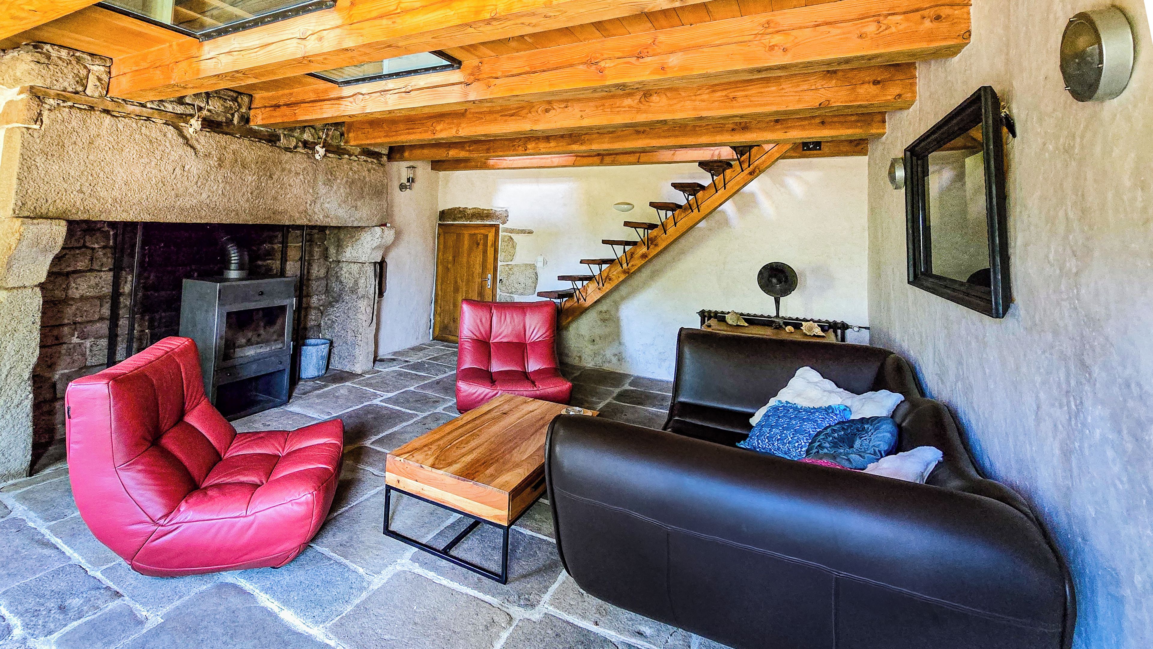 Un intérieur rustique avec une cheminée en pierre, des poutres en bois au plafond, un escalier et un coin salon agrémenté de canapés en cuir et d’un fauteuil rouge.