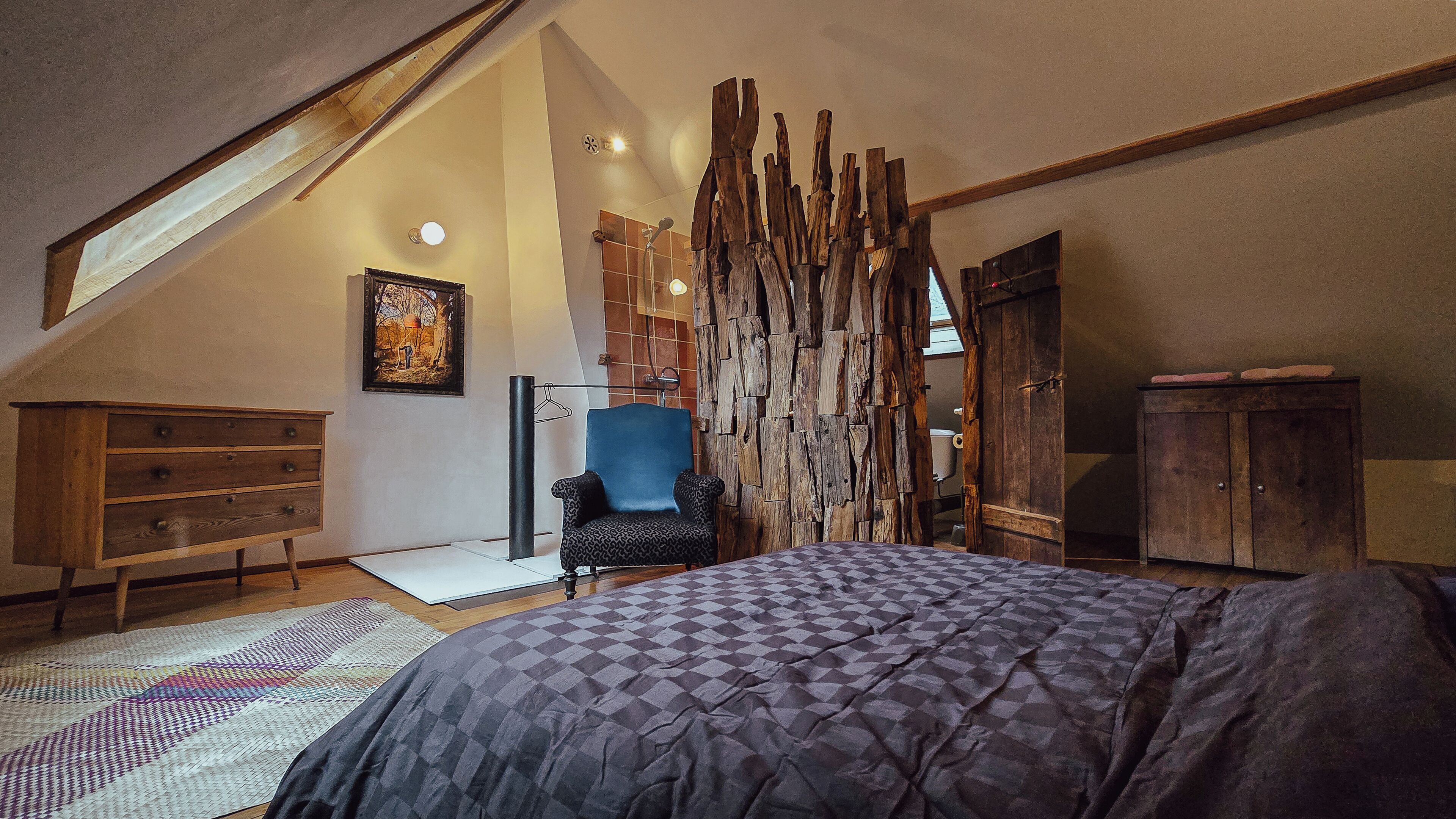 A bedroom with a sloped ceiling, a large bed, a blue chair, a wooden wardrobe, and a rustic wall panel.