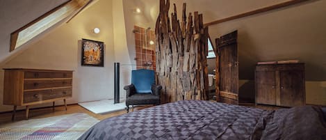 Un dormitorio con techo inclinado, una cama grande, una silla azul, un armario de madera y un panel de pared rústico.