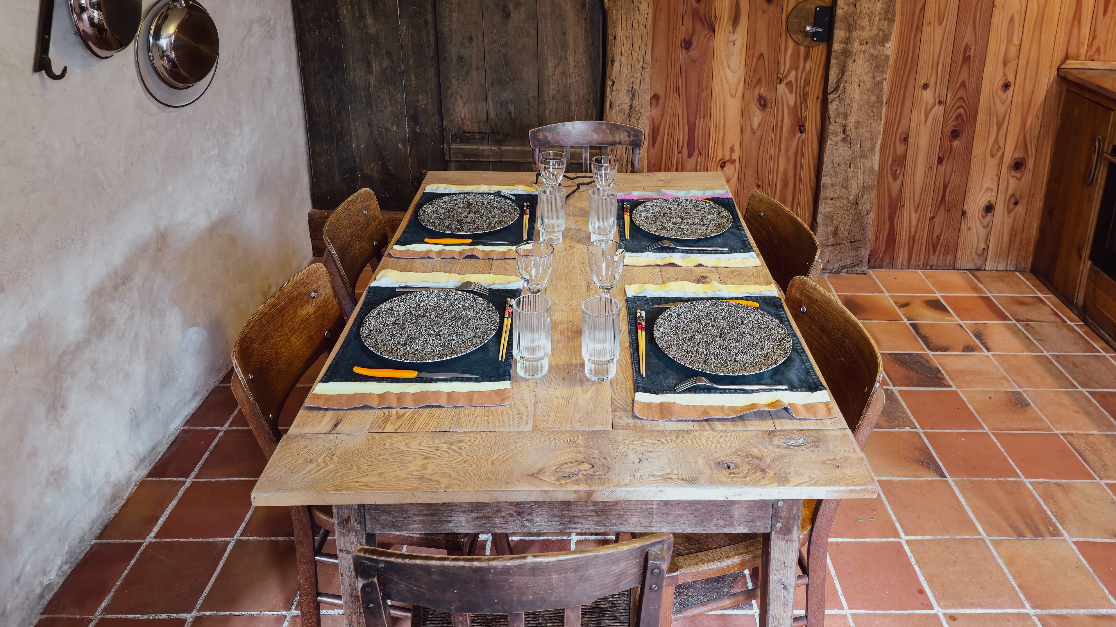 A rustic wooden dining table set with patterned plates, clear glasses, and cutlery, surrounded by wooden chairs in a room with wooden walls and tiled flooring.