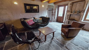 A rustic interior with a stone floor, a leather armchair, a wooden coffee table, and a stone fireplace.