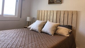 A bedroom with a wooden headboard, a quilted bedspread, two pillows, a bedside table with a lamp, and a wall-mounted display of various items. - AMSTERDAM. Exclusive Comfort on the Costa Brava (Roses)