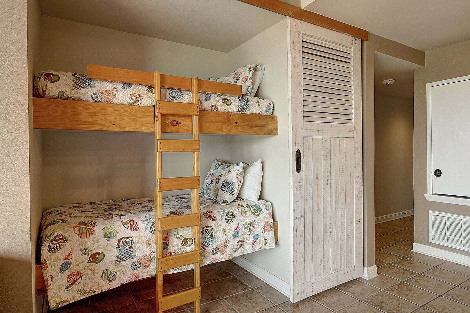 Uma cama beliche com escada de madeira, um guarda-roupa com porta veneziana e piso revestido de azulejos.