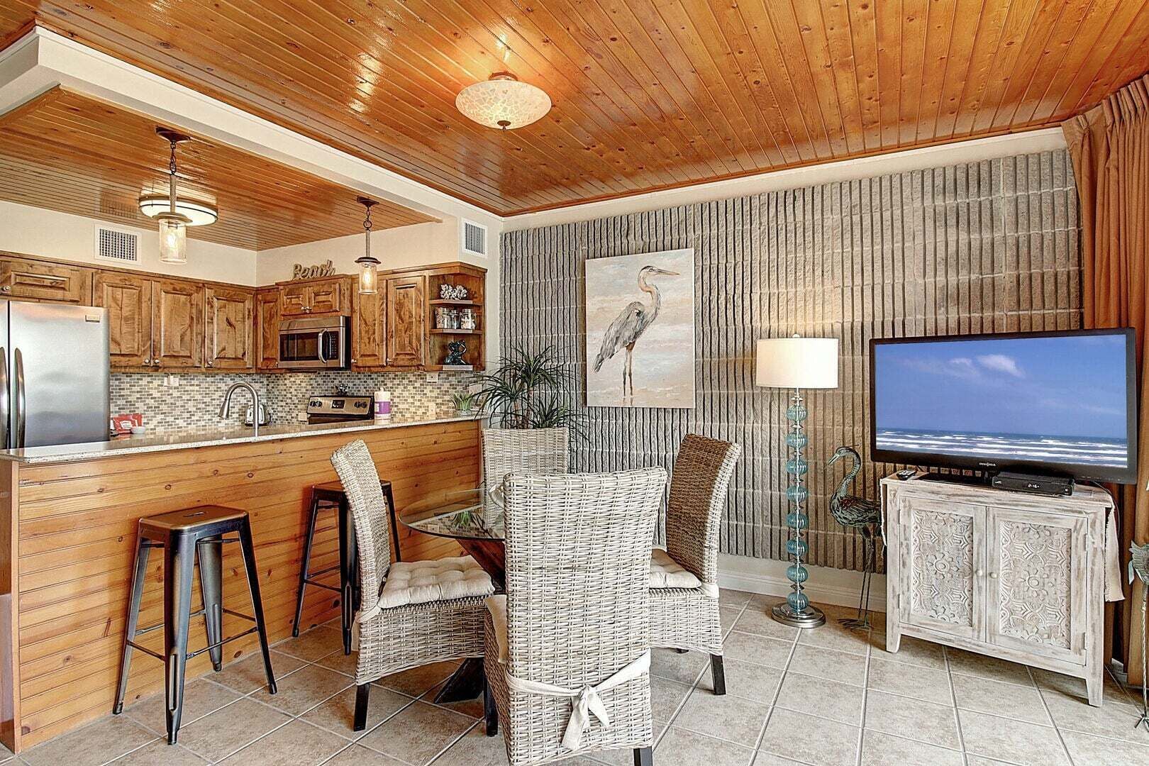 A kitchen with wooden cabinets, a microwave, and a refrigerator. There is a dining area with wicker chairs and a table. A television is on a cabinet, and a painting of a bird is on the wall.