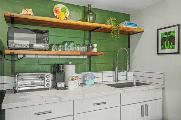 Cozinha equipada com micro-ondas, torradeira, cafeteira e pia. Prateleiras com copos e uma planta.