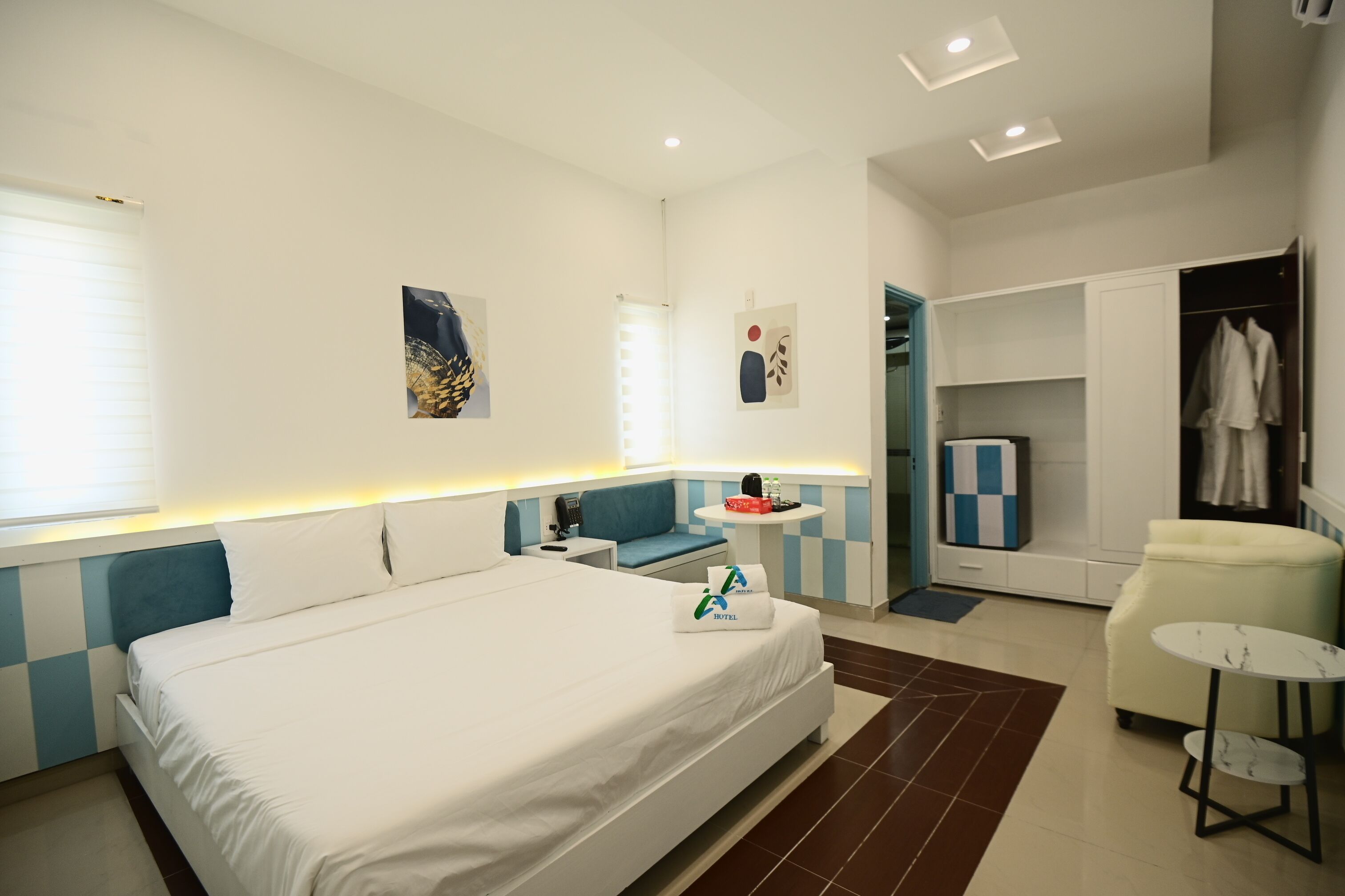 A modern hotel room with a large bed, a desk, a chair, a small table, and a wardrobe.