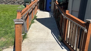 Ein Betonweg führt zu einem Gebäude mit einer blauen Tür, Holzgeländern und einer Mauer aus Stein im Hintergrund.