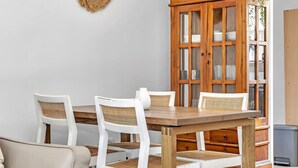 Zona pranzo con tavolo in legno e sedie bianche con sedili intrecciati, un armadietto in legno con vetrate e un oggetto decorativo a parete.