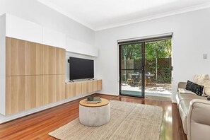 Ein modernes Wohnzimmer mit Fernseher auf einer Holzkommode, einem runden Couchtisch und einer Schiebetür zu einer Außenpatio mit Stühlen.