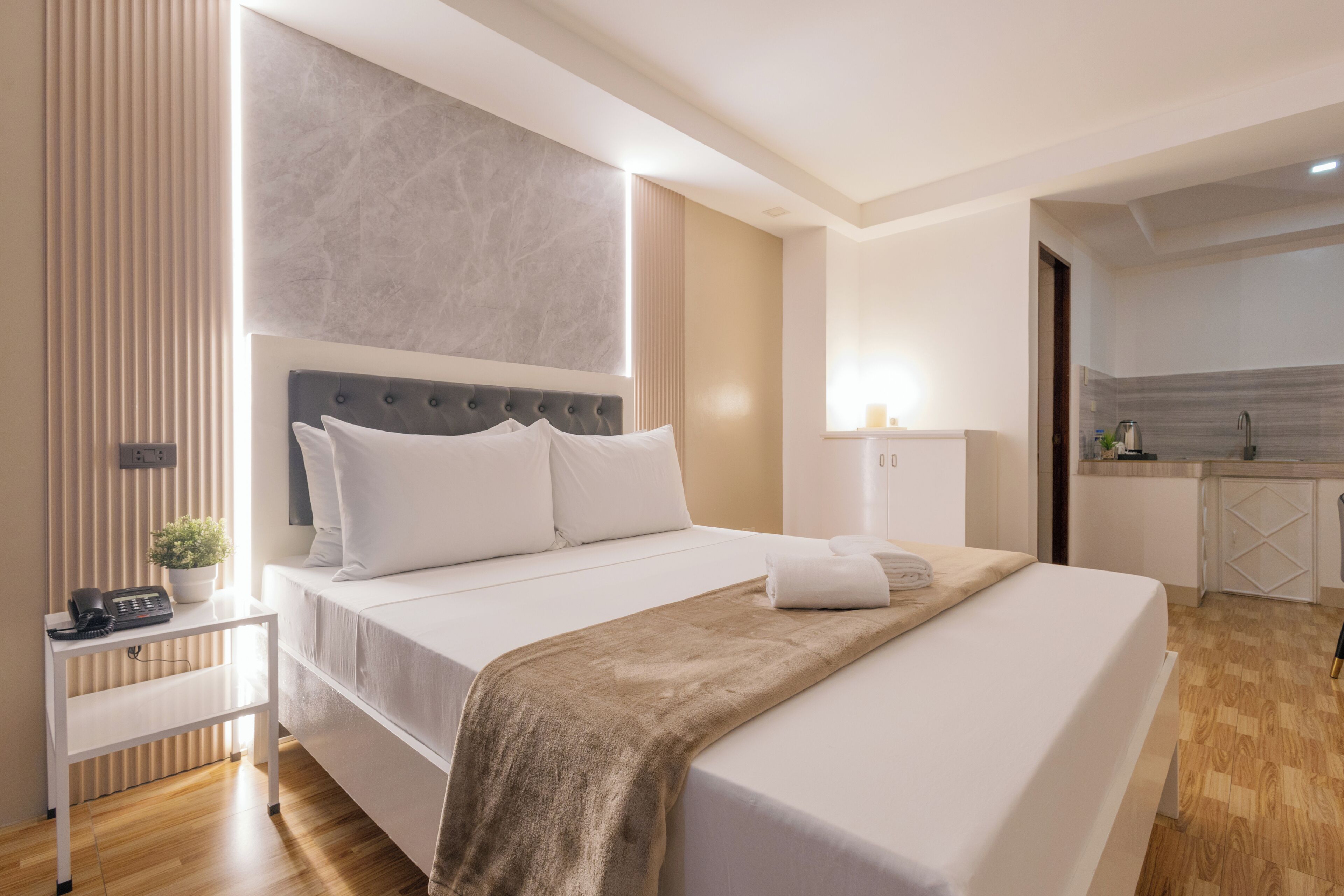 A modern hotel room with a large bed, bedside table, and a small kitchenette area.