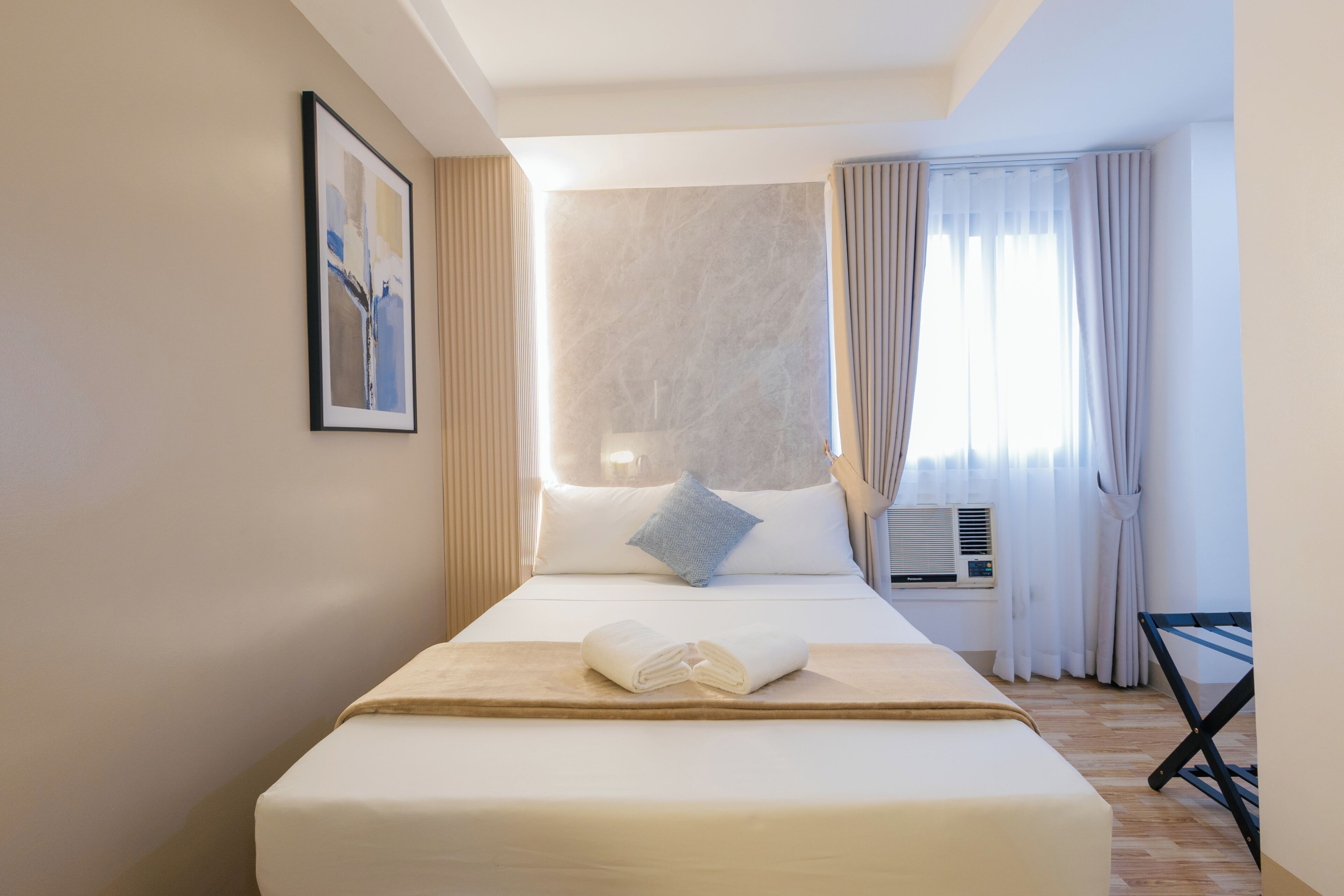 Une chambre d’hôtel avec un lit, une fenêtre avec des rideaux, un climatiseur fixé au mur et un tableau encadré accroché au mur.