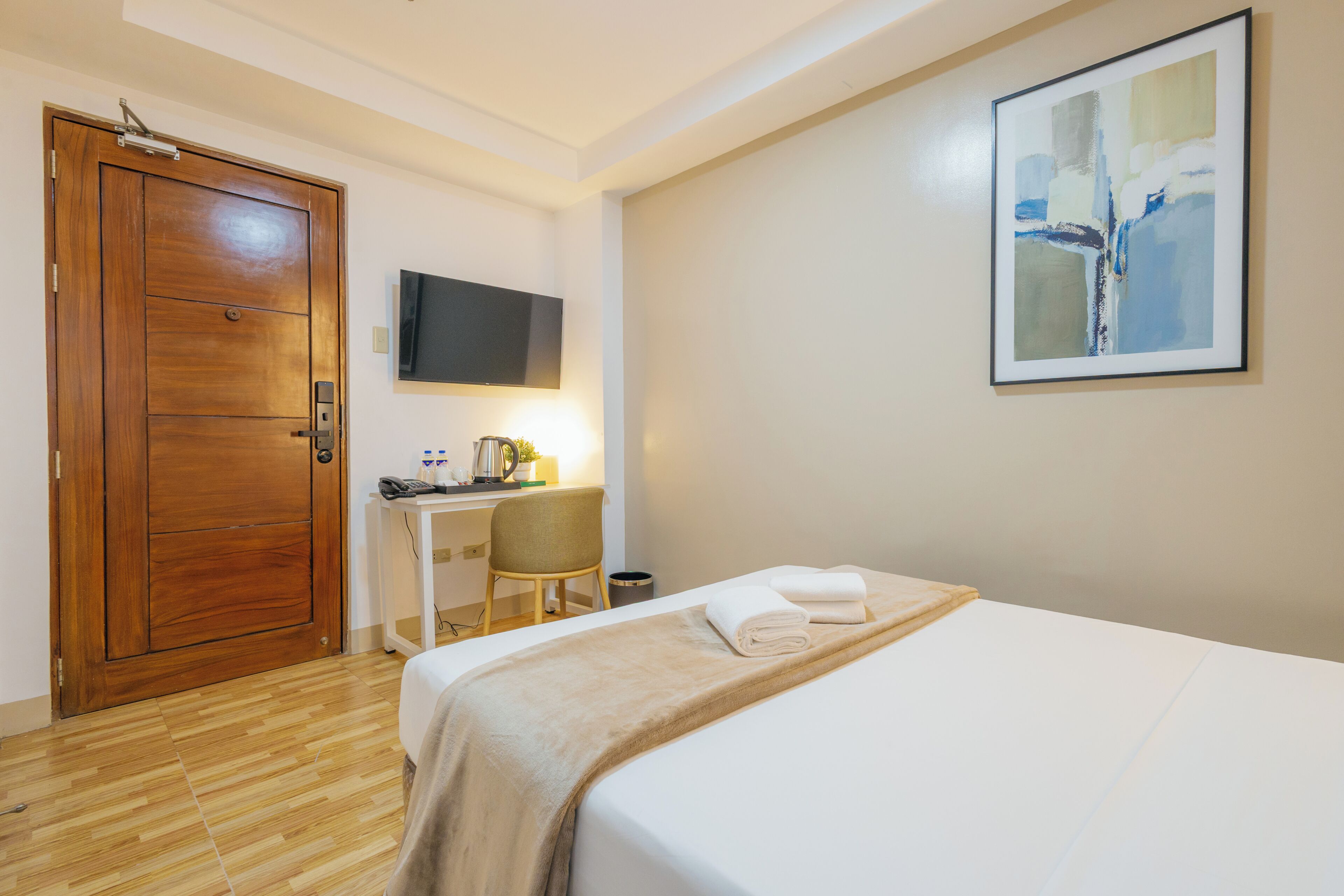 A hotel room with a wooden door, a desk with a chair, a bed with a white bedspread, and a mounted TV on the wall.