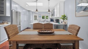Une cuisine moderne avec des meubles blancs, des appareils en acier inoxydable et un coin repas comprenant une table en bois et des chaises en osier.