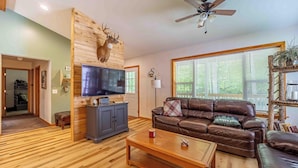 A living room with a wooden wall, a mounted deer head, a brown leather sofa, a television, and a ceiling fan.