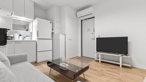 Una sala de estar moderna con una mesa de centro de vidrio, sofá gris, televisor de pantalla plana, armarios blancos y una cocina con refrigerador y microondas.