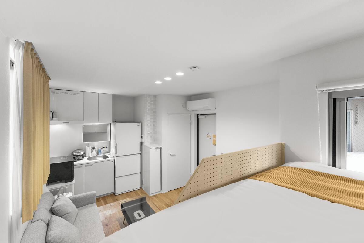 A compact hotel room with a kitchenette, a bed with a patterned headboard, and a window with blinds.