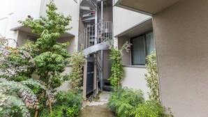 Un patio estrecho con una escalera espiral que conduce a un piso superior, rodeado de vegetación y fachada de edificio.
