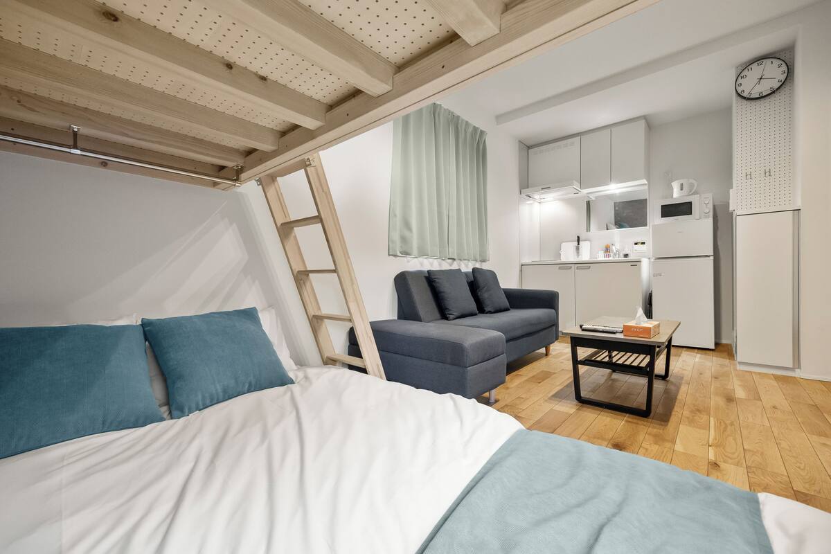 A bedroom with a bed, a sofa, a small table, a ladder, a kitchenette, and a clock on the wall.