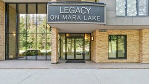 The exterior of a building labeled "Legacy on Mara Lake" with a glass entrance, stone facade, and a clear view of the surrounding trees.