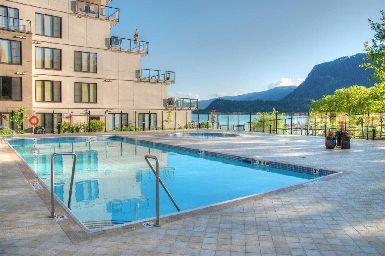 A swimming pool with a diving area, surrounded by a building with balconies and a view of a lake and mountains.
