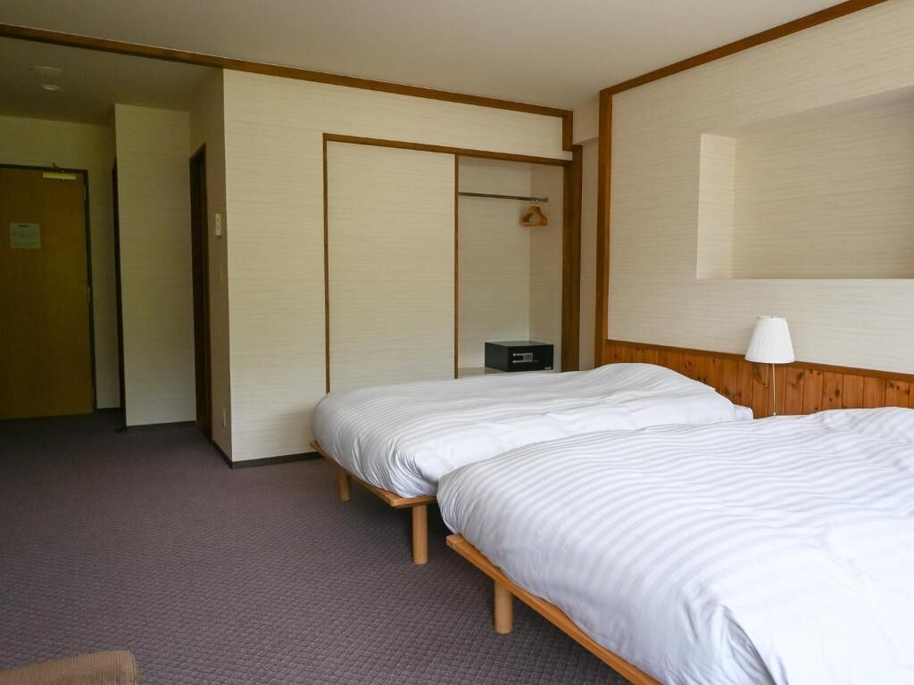 A hotel room with two beds, a wooden headboard, a bedside table, a lamp, and a wardrobe with a mirror.