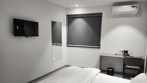 Et hotelværelse med en seng, et skrivebord, en stol, et fjernsyn, et spejl og et klimaanlæg.