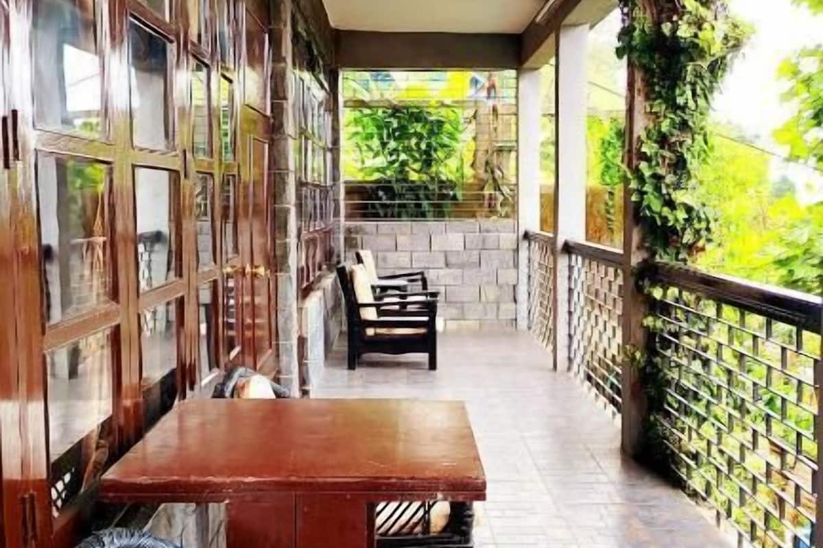 A covered patio with a wooden table and wicker chairs, a stone wall, and a view of greenery.