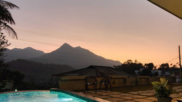 A swimming pool with clear water, surrounded by a tiled deck and a potted plant. In the background, there is a mountain and a building with a sloped roof. - Maison Claire Service Villa Wayanad – 4BR Stay Near Attractions (Kottappadi part, Wayand)